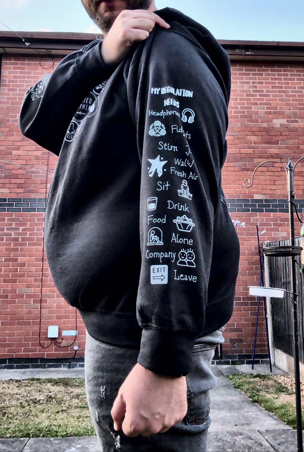 A man wearing a black hoodie with white graphic text and icons printed down the left sleeve, standing outdoors in front of a red brick wall.