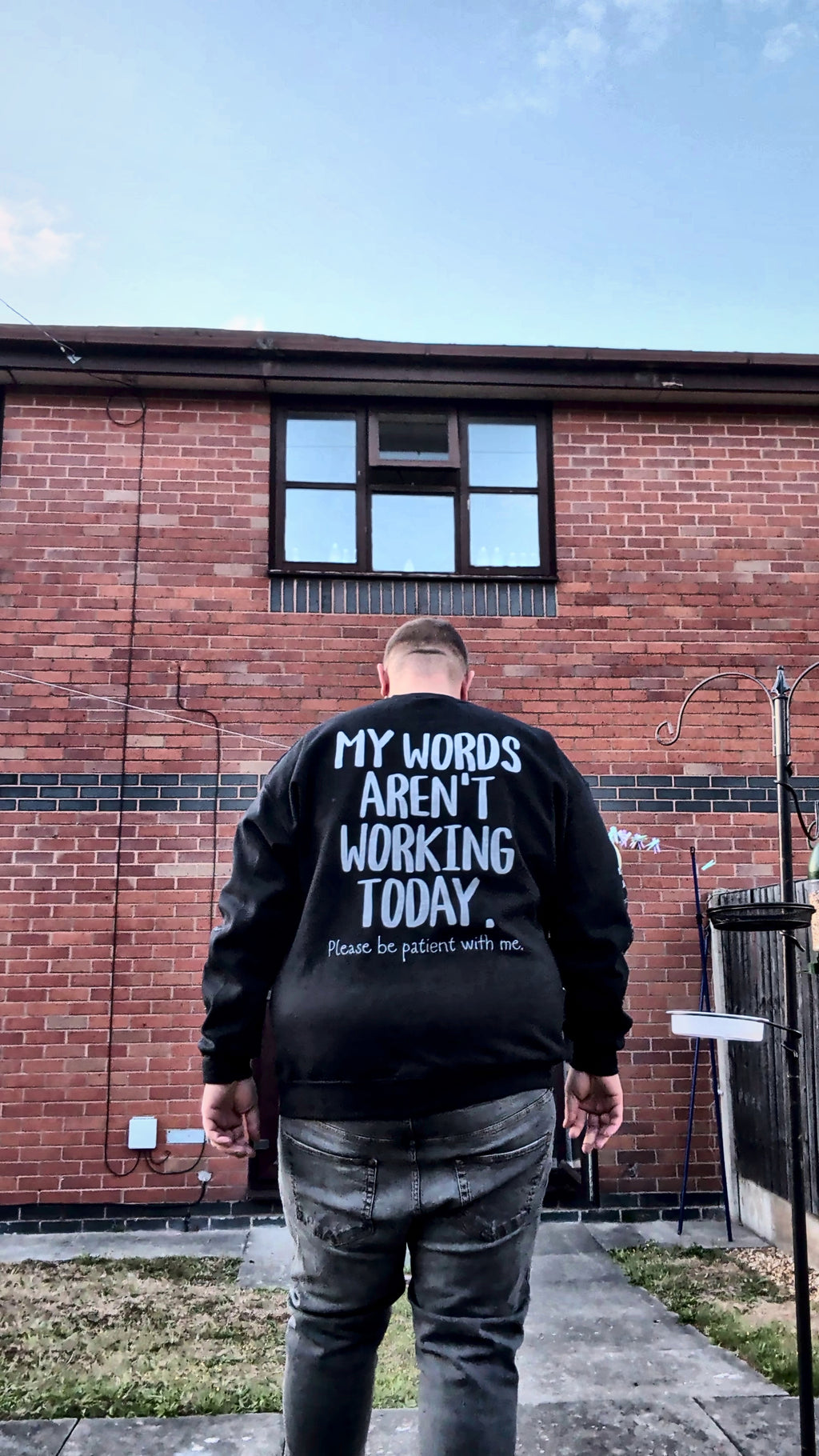 A man viewed from behind wearing a black hoodie with white text that reads 'MY WORDS AREN'T WORKING TODAY.' standing outdoors in front of a red brick building.