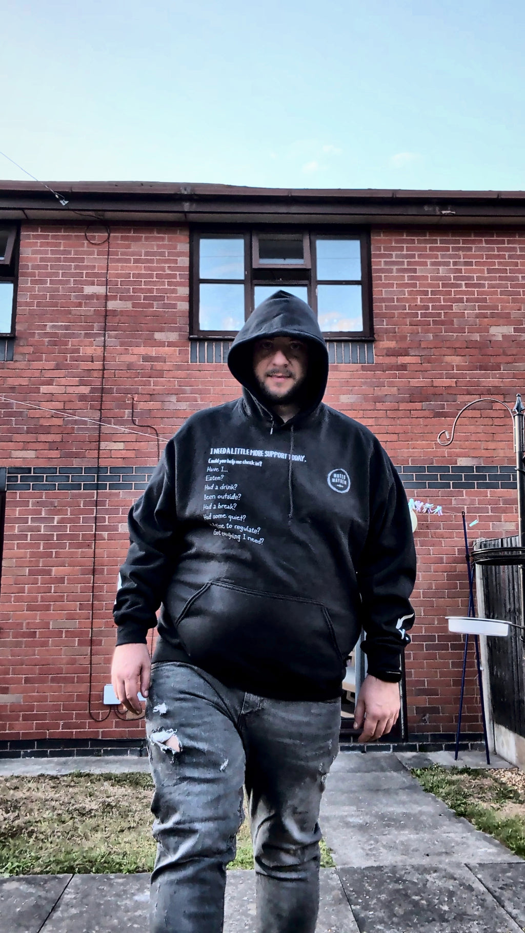 A man wearing a black hoodie with white text and distressed gray jeans, standing in front of a red brick house.