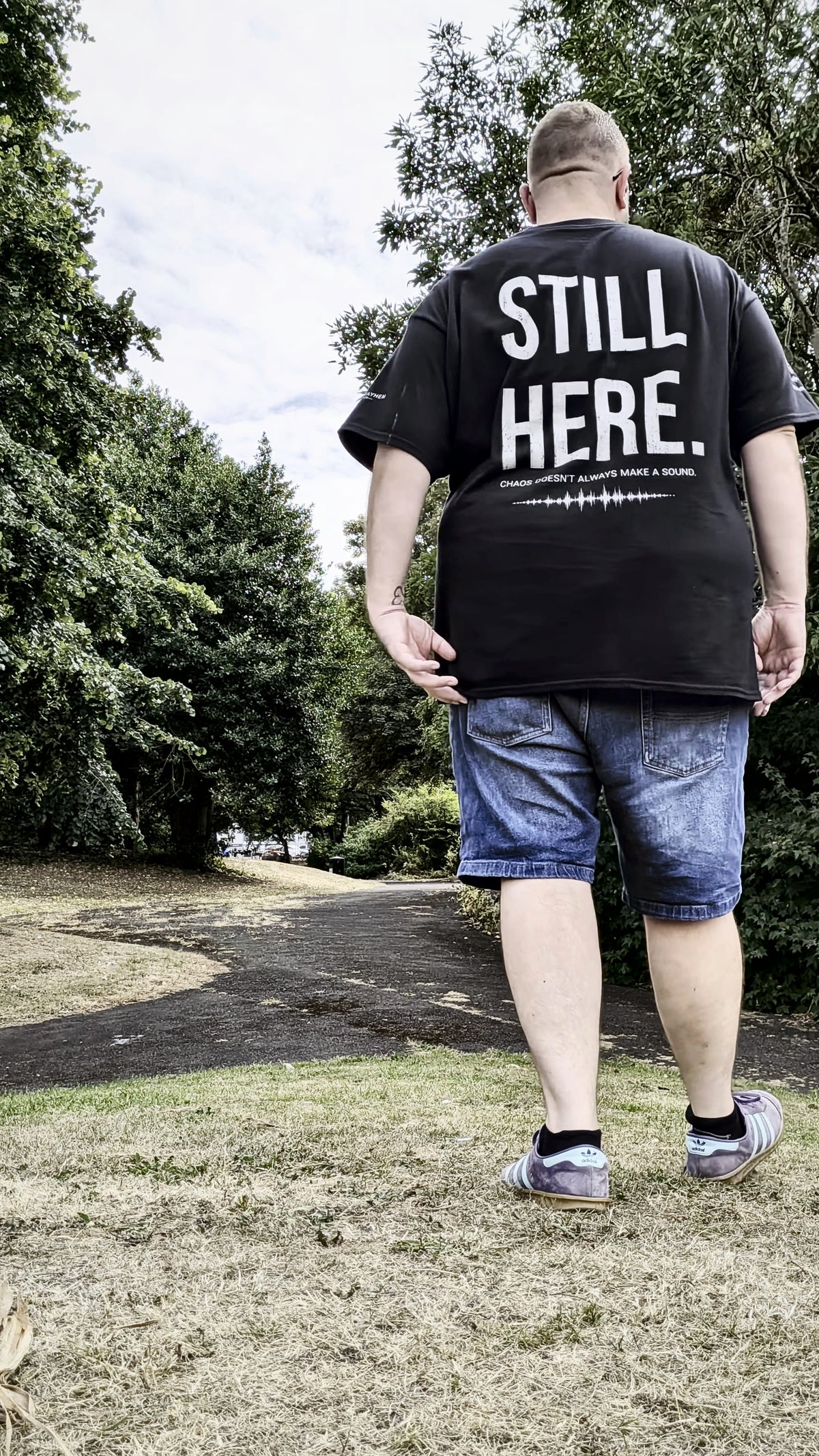 A man viewed from behind wearing a black t-shirt with the text 'STILL HERE.' printed in white on the back, blue denim shorts, and sneakers, walking on a grassy path surrounded by trees.