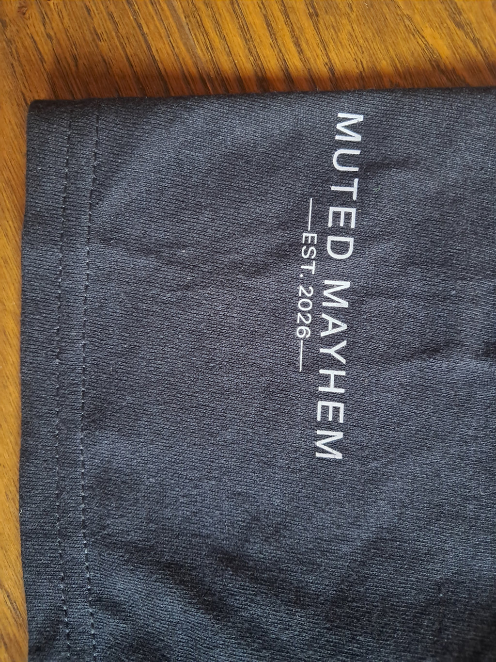 Close-up of a dark charcoal gray fabric item, likely a t-shirt sleeve, featuring vertical white text that reads 'MUTED MAYHEM EST. 2026' on a wooden surface promoting mental health and neurodivergence awareness by muted mayhem.