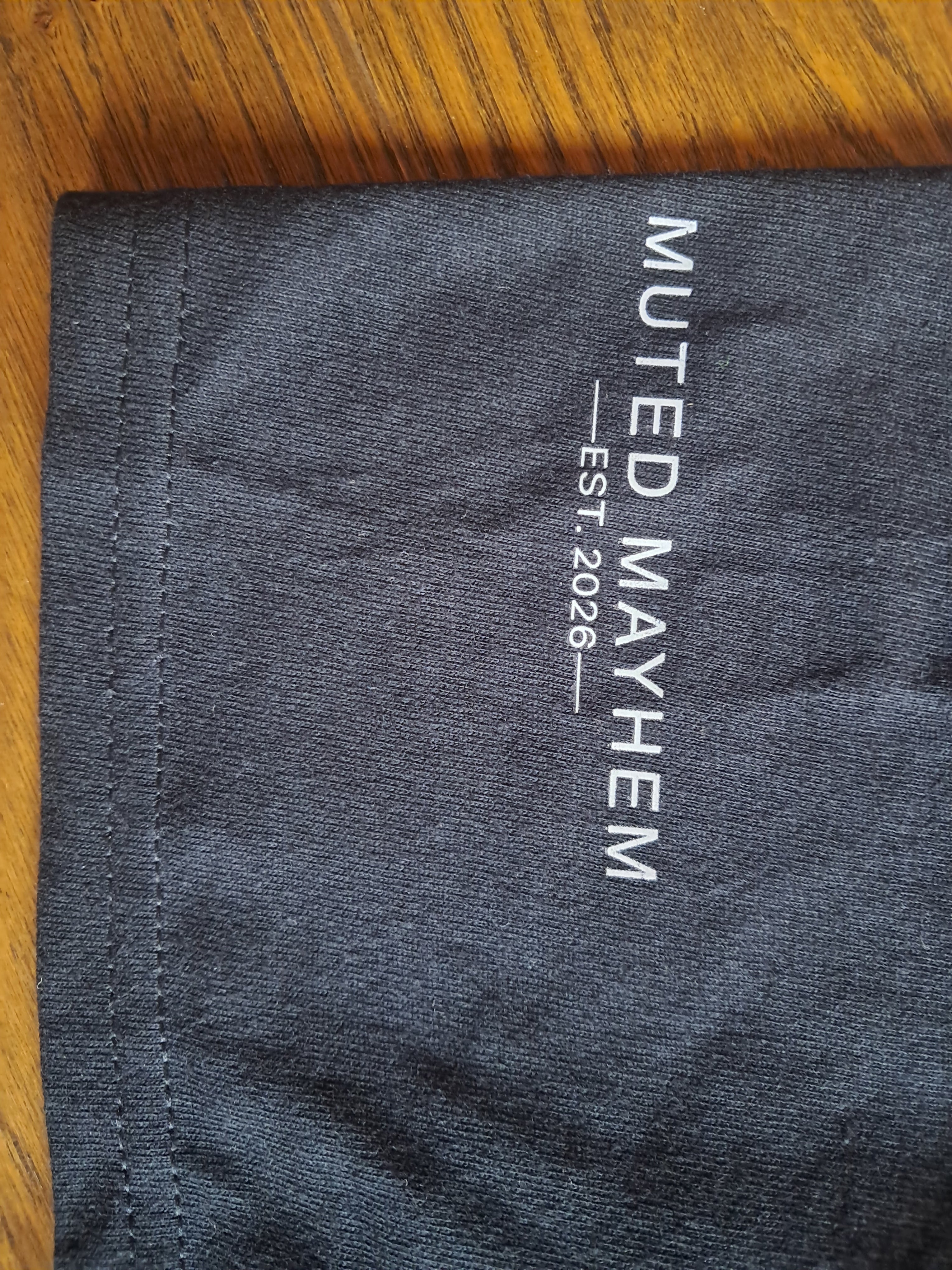 Close-up of a dark charcoal gray fabric item, likely a t-shirt sleeve, featuring vertical white text that reads 'MUTED MAYHEM EST. 2026' on a wooden surface promoting mental health and neurodivergence awareness by muted mayhem.