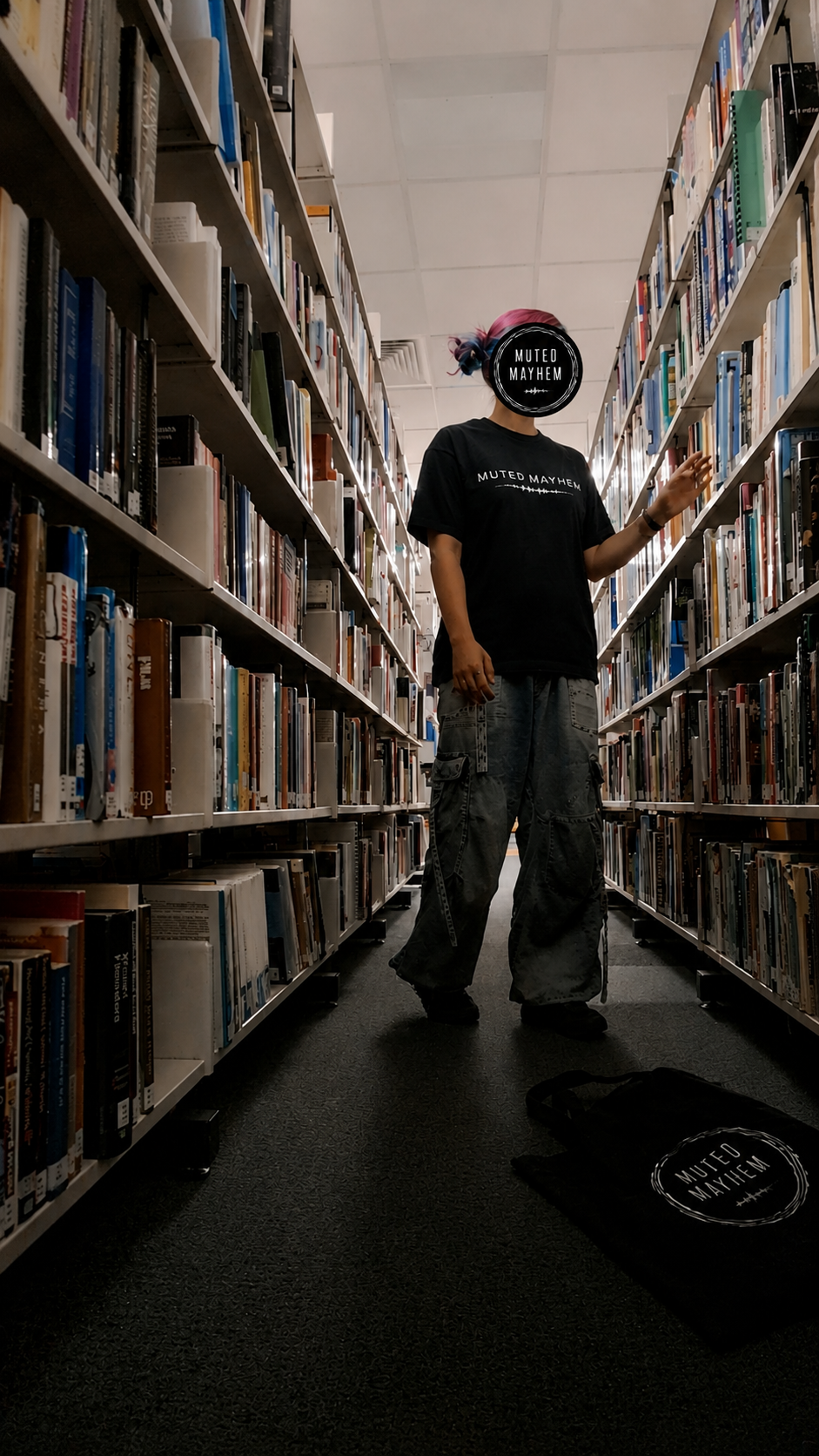 A person standing in a library aisle between tall bookshelves, wearing a black t-shirt, gray jeans. A black tote bag with the same logo is on the floor nearby promoting mental health and neurodivergence awareness by muted mayhem.