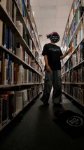 A person standing in a library aisle between tall bookshelves, wearing a black t-shirt, gray jeans. A black tote bag with the same logo is on the floor nearby promoting mental health and neurodivergence awareness by muted mayhem.