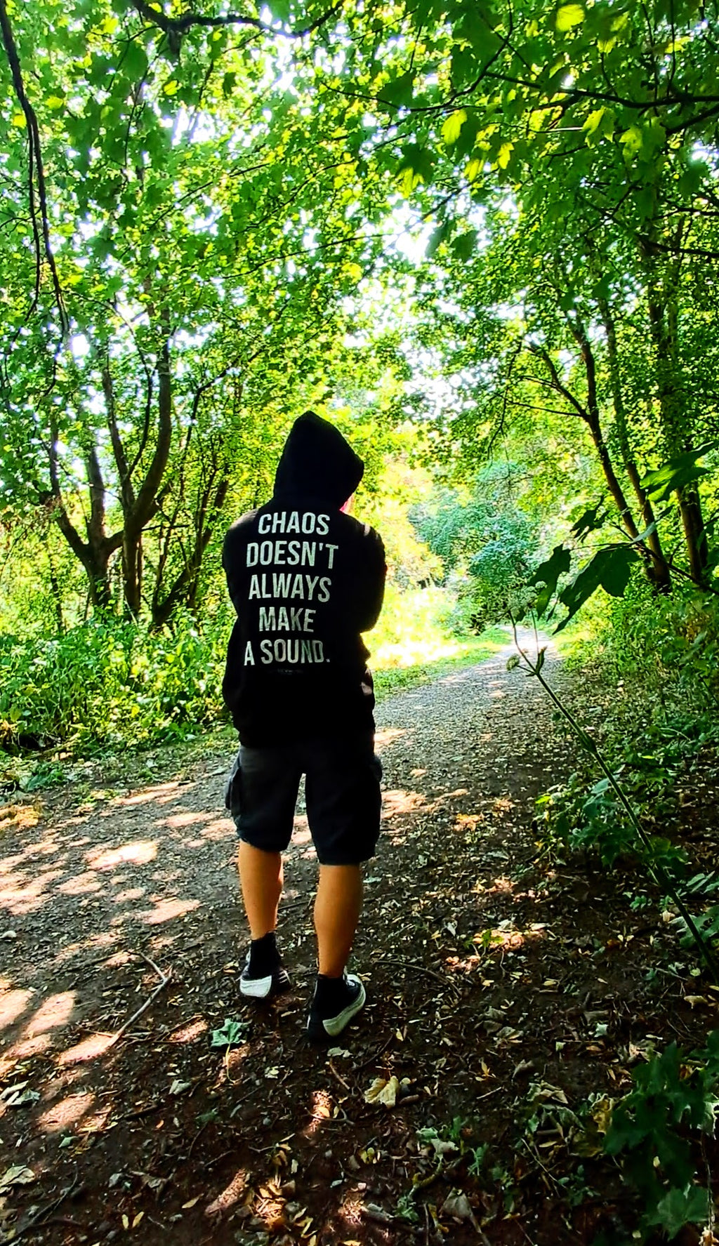 A person viewed from behind wearing a black hoodie with the white text 'CHAOS DOESN'T ALWAYS MAKE A SOUND.' and black shorts, walking on a sun-dappled forest path promoting mental health and autism and adhd awareness.