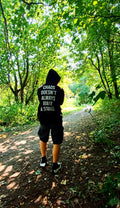 A person viewed from behind wearing a black hoodie with the white text 'CHAOS DOESN'T ALWAYS MAKE A SOUND.' and black shorts, walking on a sun-dappled forest path promoting mental health and autism and adhd awareness.