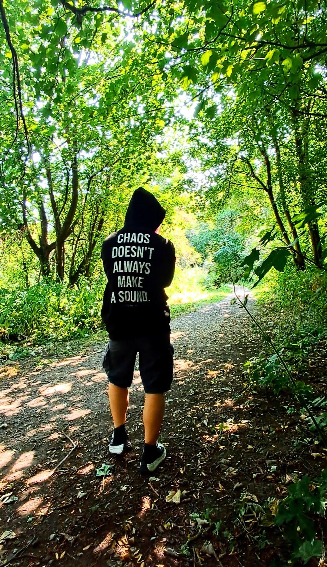 A person viewed from behind wearing a black hoodie with the white text 'CHAOS DOESN'T ALWAYS MAKE A SOUND.' and black shorts, walking on a sun-dappled forest path promoting mental health and autism and adhd awareness.