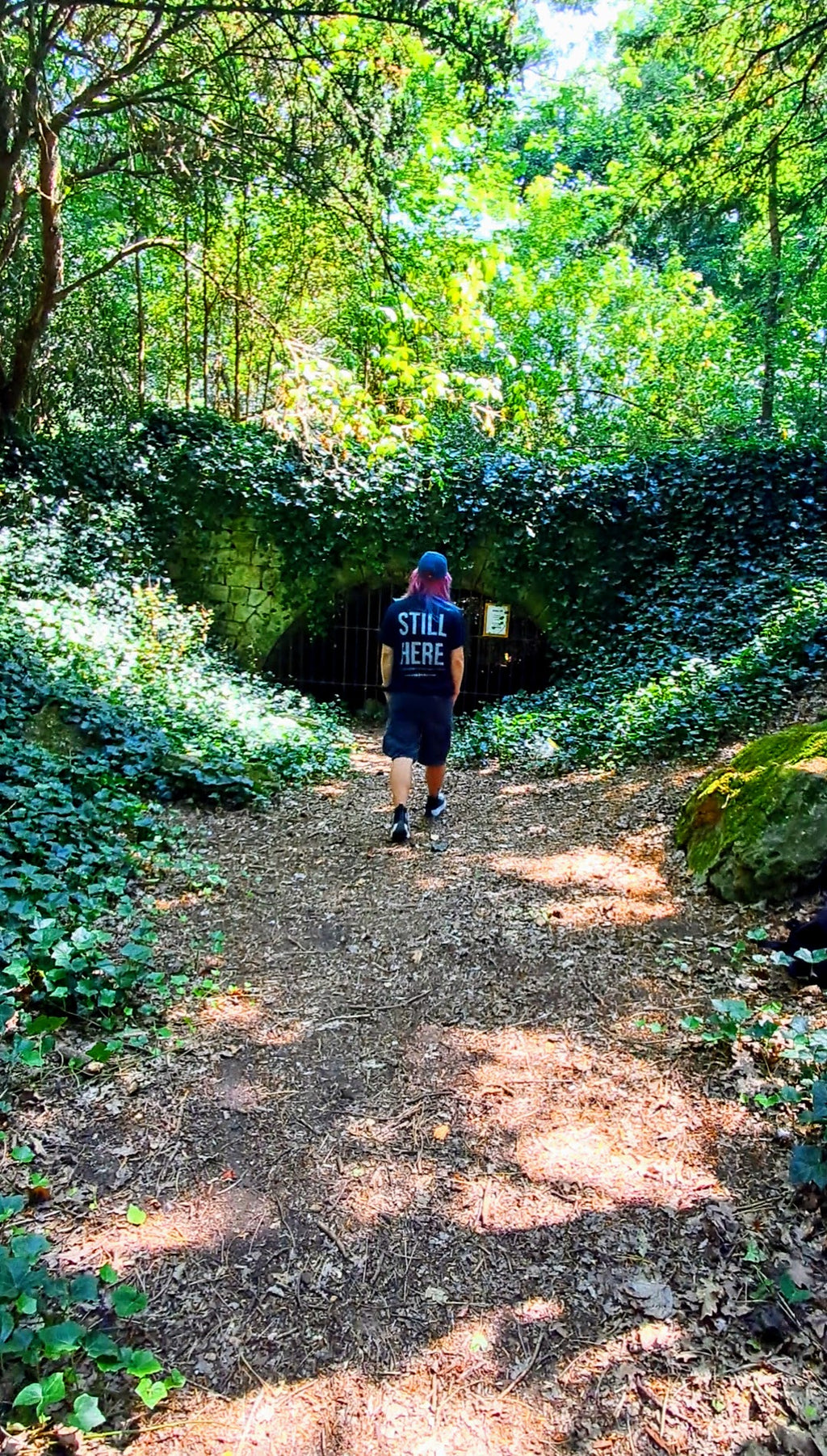 A person wearing a black t-shirt with 'STILL HERE' printed on the back and black shorts walks away on a dirt path surrounded by lush green forest foliage. Promoting mental health, autism and ADHD awareness