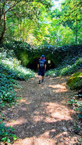 A person wearing a black t-shirt with 'STILL HERE' printed on the back and black shorts walks away on a dirt path surrounded by lush green forest foliage. Promoting mental health, autism and ADHD awareness