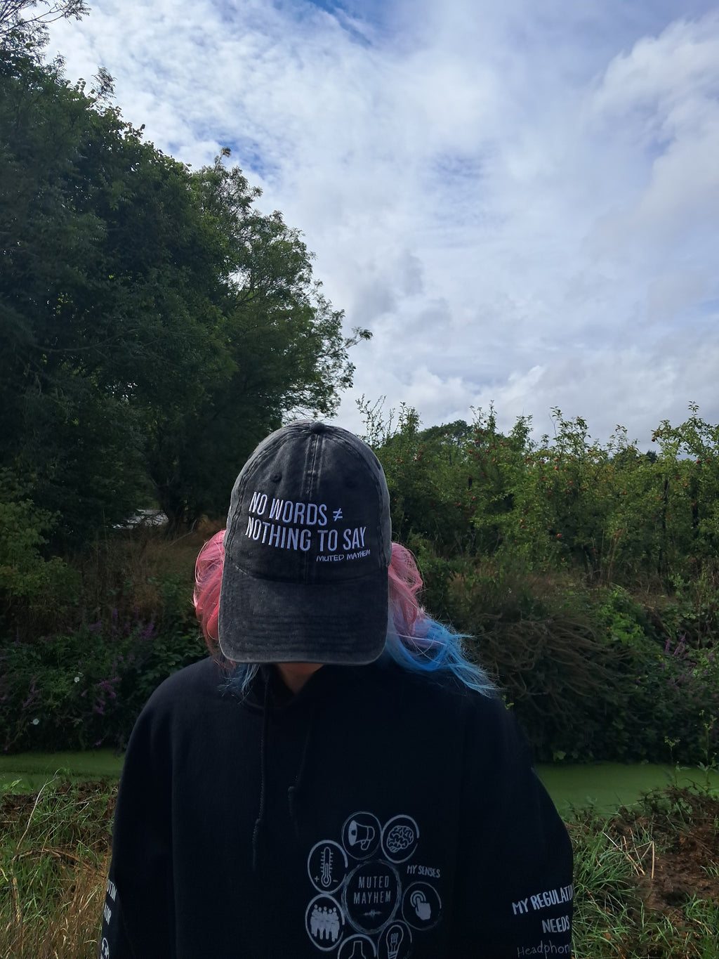 A person outdoors wearing a dark gray baseball cap with the text 'NO WORDS = NOTHING TO SAY' and a black hoodie, with pink and blue hair visible, set against a background of trees and a cloudy sky.