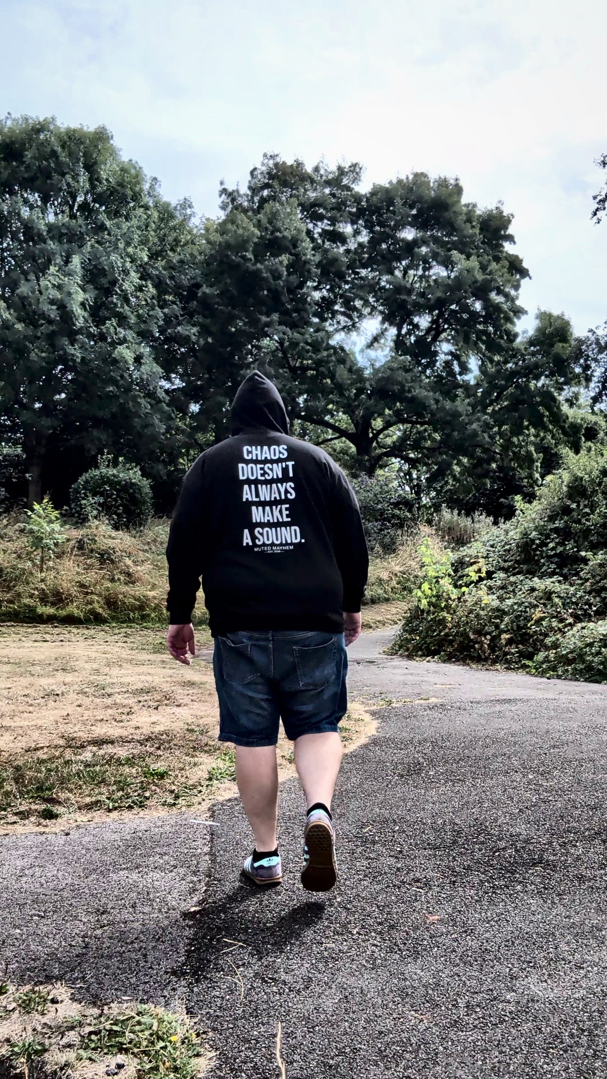 A man is seen from behind walking on a paved path outdoors, wearing a black hoodie with white text on the back that reads 'CHAOS DOESN'T ALWAYS MAKE A SOUND.' and dark denim shorts.