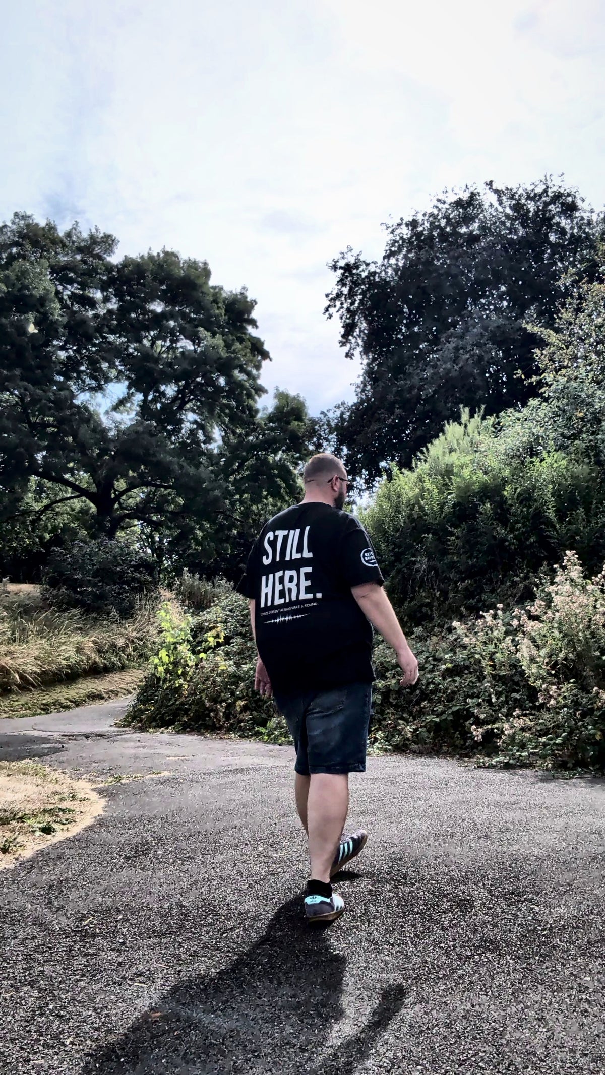 A man walking away on a paved path surrounded by trees and bushes, wearing a black t-shirt with 'STILL HERE.' printed in white on the back.
