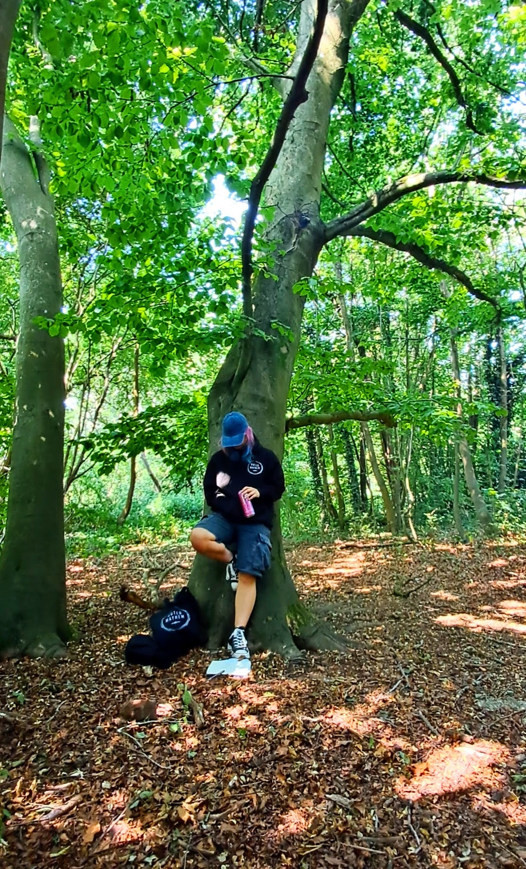 A person sitting on the base of a large tree in a wooded forest, wearing a black hoodie, blue cap, and shorts, with a black backpack on the ground nearby.