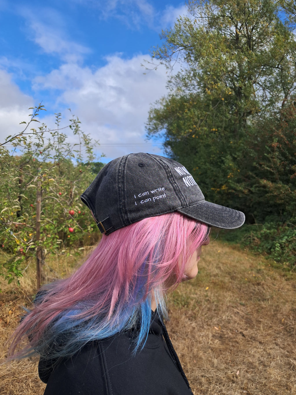 A person outdoors wearing a black baseball cap with white embroidered text and long pink hair with blue tips, standing in a grassy field with trees and a blue sky in the background.