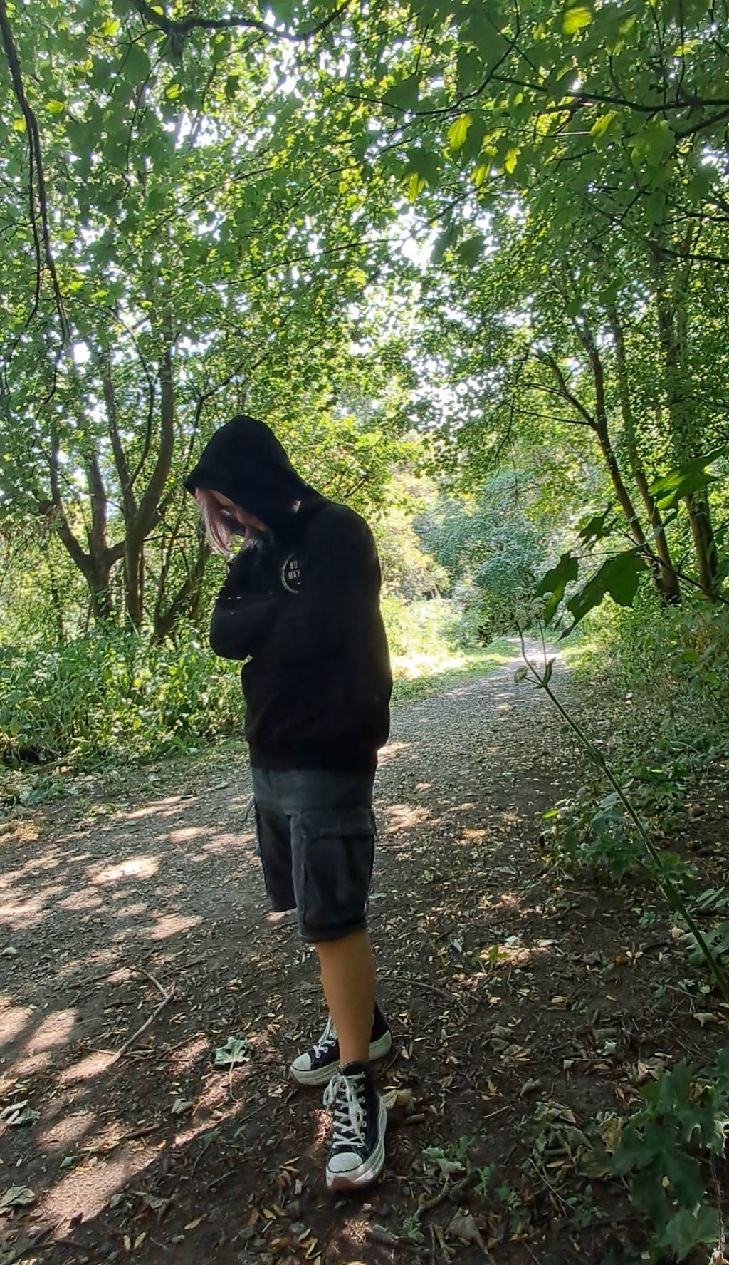 A person wearing a black hoodie, dark gray shorts, and white sneakers stands on a dirt path in a wooded area with trees and foliage promoting mental health, autism and adhd awareness 