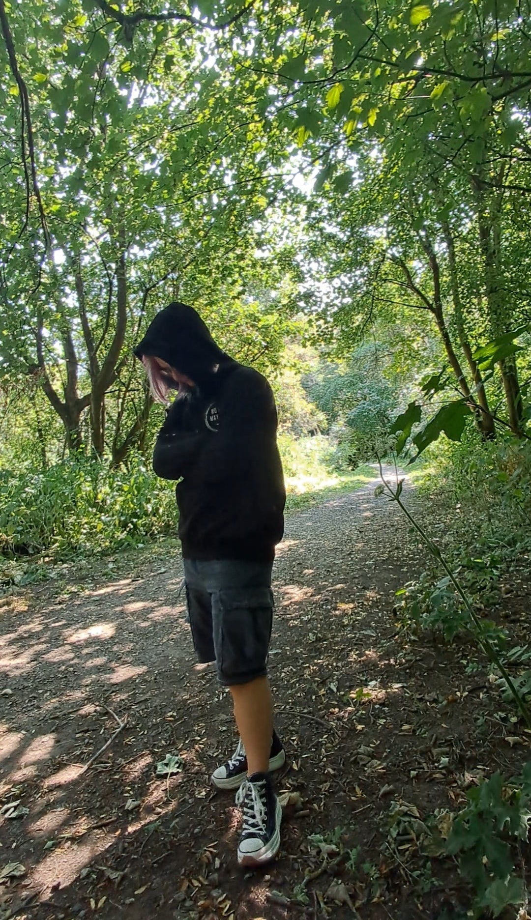 A person wearing a black hoodie, dark gray shorts, and white sneakers stands on a dirt path in a wooded area with trees and foliage promoting mental health, autism and adhd awareness 
