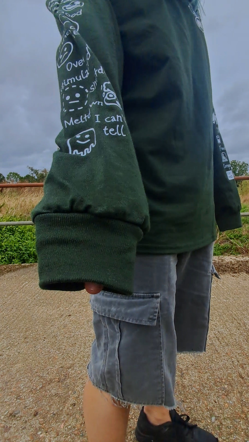 A person outdoors wearing a dark green long-sleeve sweatshirt with white graphic text and smiley face prints on the sleeves, paired with gray cargo shorts and black sneakers.