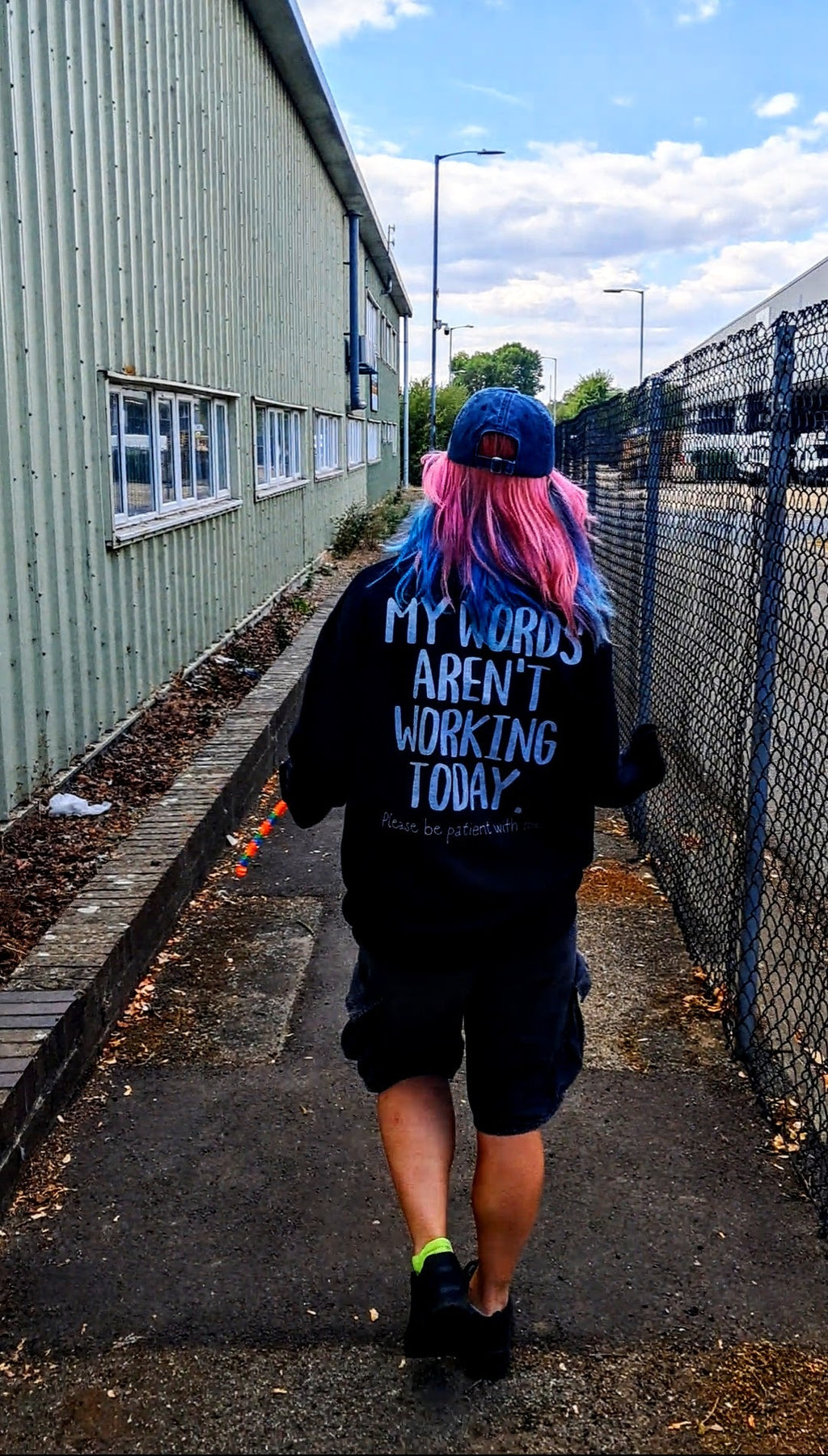 A person viewed from behind walking down an outdoor path, wearing a black hoodie with the text 'MY WORDS AREN'T WORKING TODAY.' in blue, black shorts, and a blue cap.