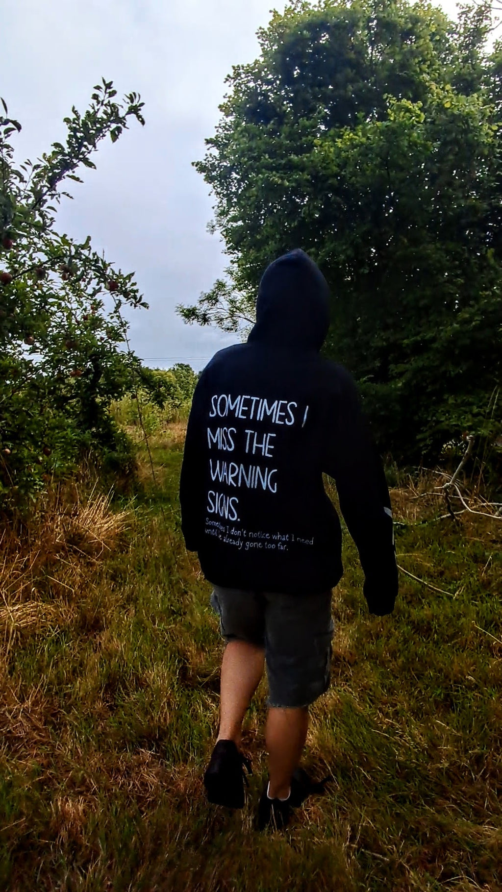 A person viewed from behind walking on a grassy path outdoors, wearing a black hoodie with white text on the back that reads, 'SOMETIMES I MISS THE WARNING SIGNS.'.