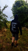 A person viewed from behind walking on a grassy path outdoors, wearing a black hoodie with white text on the back that reads, 'SOMETIMES I MISS THE WARNING SIGNS.'.