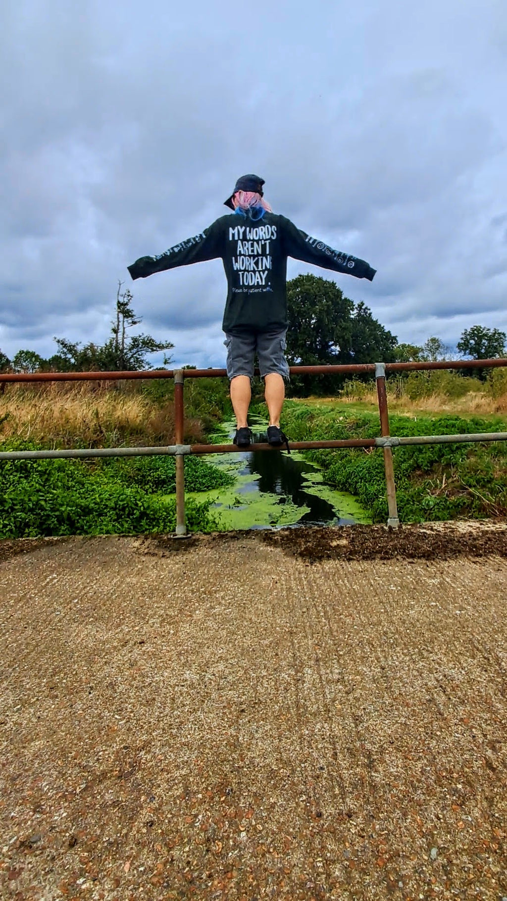 A person stands on a metal railing outdoors with arms outstretched, wearing a black long-sleeve shirt with white text that reads 'MY WORD ARE NOT WORKING TODAY', gray shorts, and a black cap. The background shows grass, trees, and a cloudy sky, with a gravel path in the foreground.