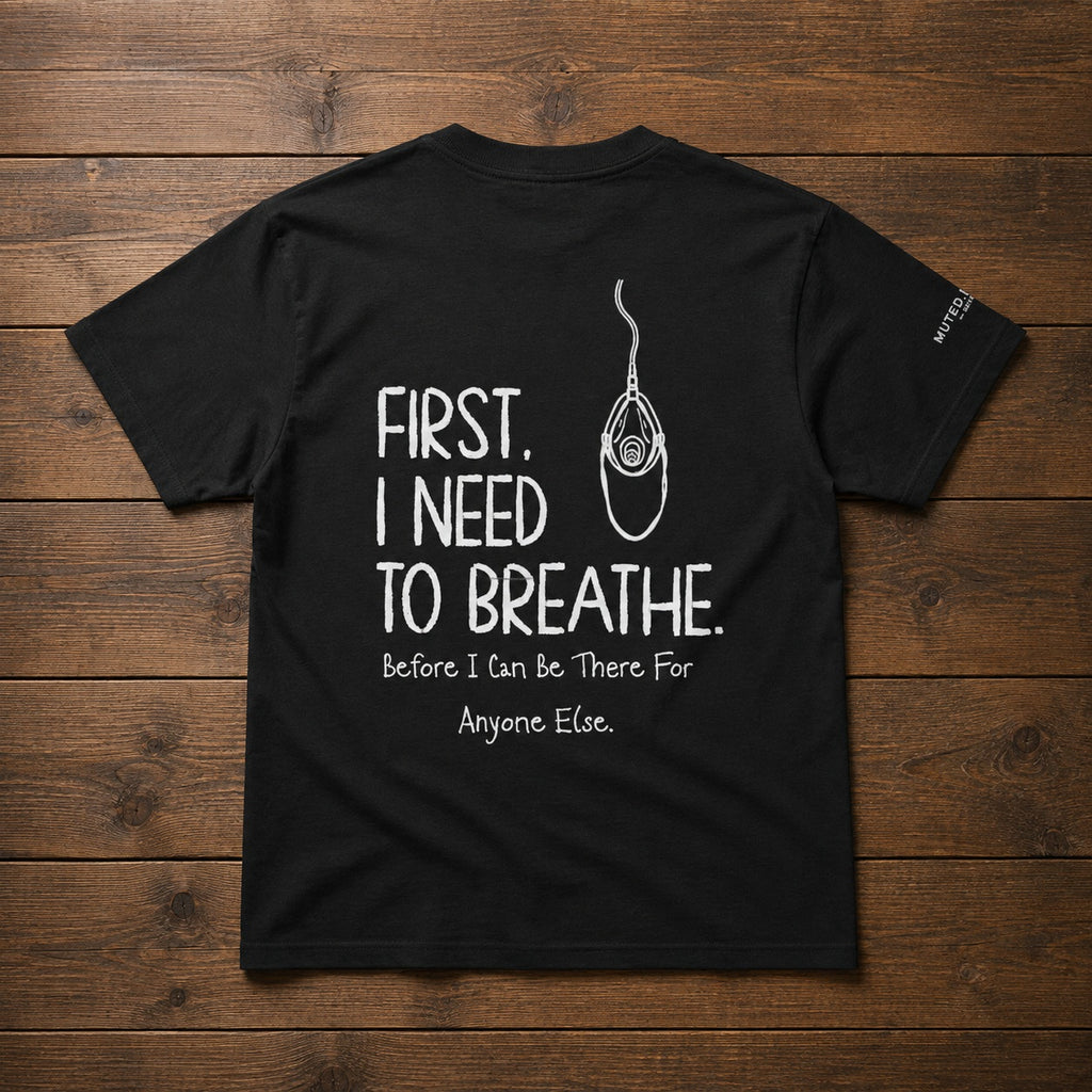 A black short-sleeve t-shirt laid flat on a dark wood plank background, featuring a white graphic print on the back with the text 'FIRST, I NEED TO BREATHE. Before I can be There For Anyone Else.