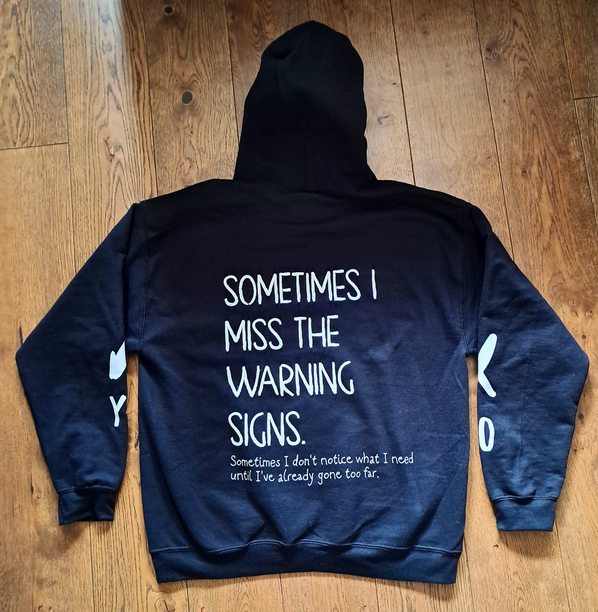 A dark navy blue hoodie is laid flat on a wooden floor, showing the back side with white text that reads 'SOMETIMES I MISS THE WARNING SIGNS.' and smaller text below.