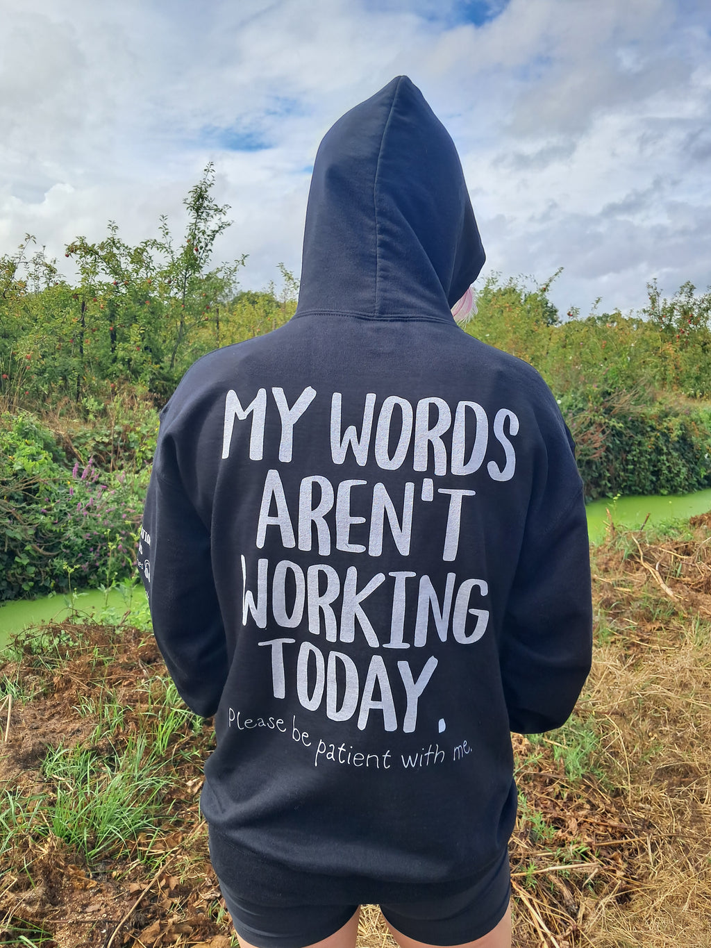A person standing outdoors in a field, viewed from behind, wearing a dark navy hoodie with large white text on the back that reads, 'MY WORDS AREN'T WORKING TODAY.' with smaller text below, paired with black shorts.