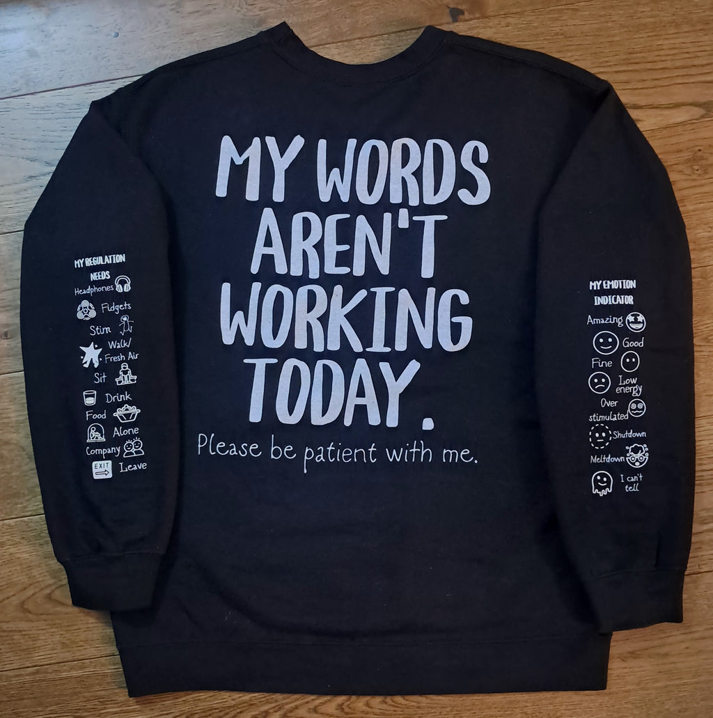 A black long-sleeve sweatshirt laid flat on a wooden surface, showing the back side with large white text that reads 'MY WORDS AREN'T WORKING TODAY.' and smaller text below reading 'Please be patient with me.