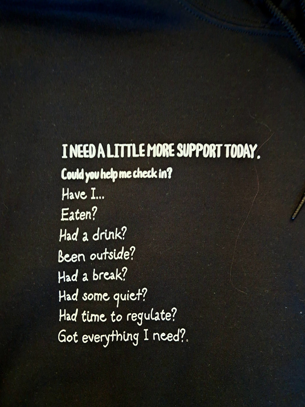 A close-up of a black garment, likely a hoodie or sweatshirt, featuring white printed text that reads: 'I NEED A LITTLE MORE SUPPORT TODAY. Could you help me check in? Have I.