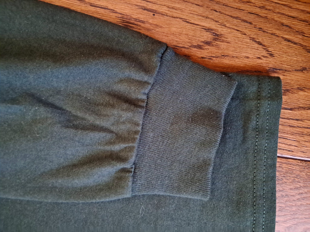 Close-up of an olive green garment sleeve cuff with a gathered elastic seam, laid flat on a wooden surface.