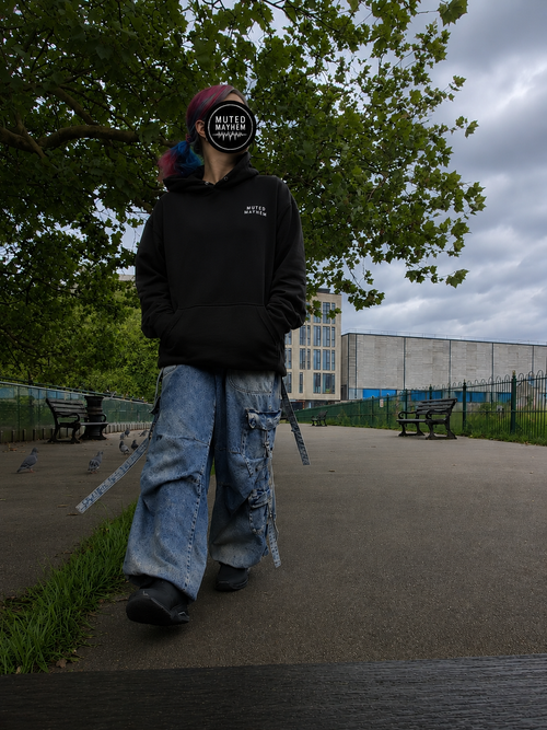 A person walking outdoors in a park wearing a black hoodie with a small white logo on the chest, distressed light blue jeans, promoting mental health and neurodivergence awareness by muted mayhem.