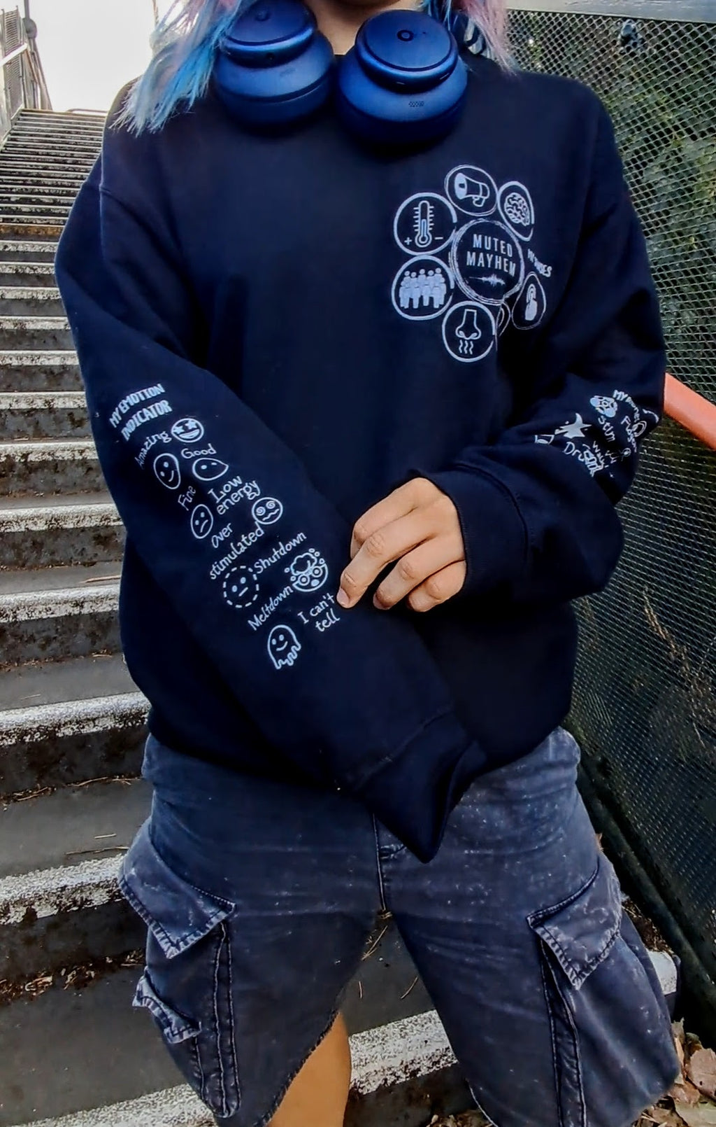A person wearing a black sweatshirt with white graphic prints on the chest and sleeves, paired with dark cargo shorts and blue headphones around the neck, standing on outdoor concrete stairs.