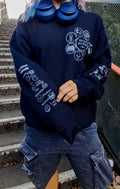 A person wearing a black sweatshirt with white graphic prints on the chest and sleeves, paired with dark cargo shorts and blue headphones around the neck, standing on outdoor concrete stairs.