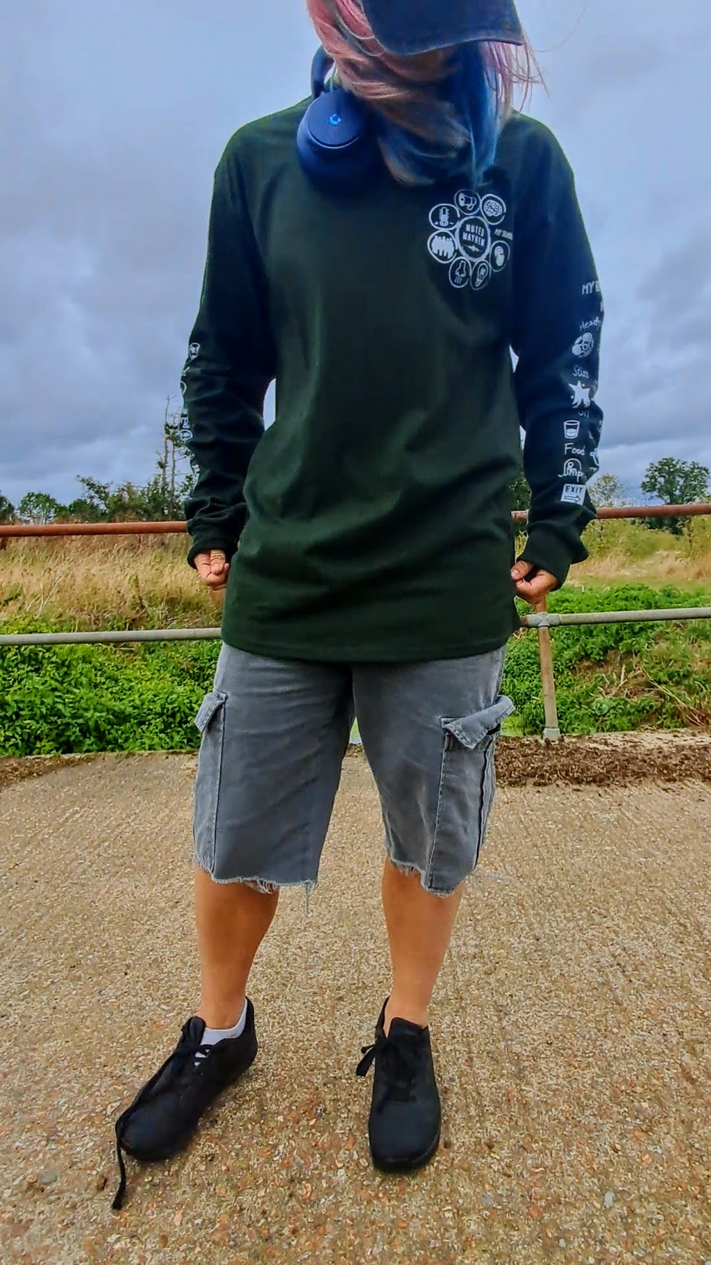 A person standing outdoors wearing a dark green long-sleeve graphic shirt, gray cargo shorts, and black sneakers, with a blue face mask hanging around the neck and a cap on.