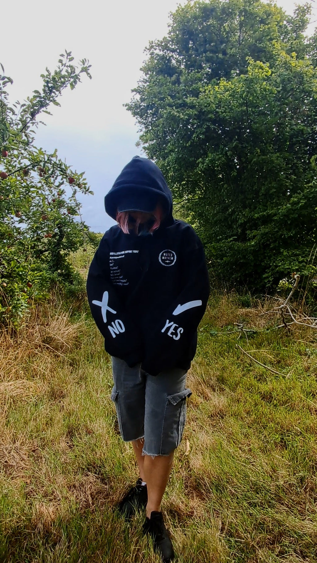A person wearing a black hoodie with white graphic text and logos, gray cargo shorts, and black sneakers, standing outdoors in a grassy field with trees in the background.