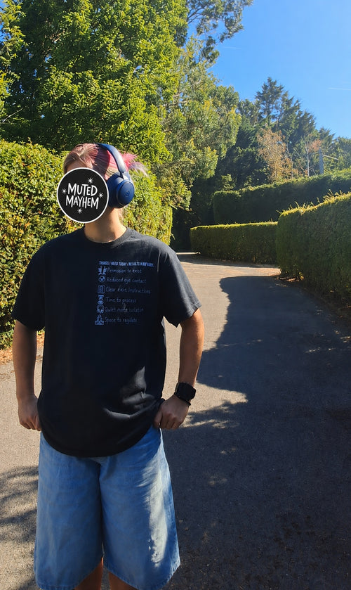 A person standing outdoors on a paved path wearing a black graphic t-shirt and light blue denim shorts. The person is wearing blue over-ear headphones and a black circular sticker with the text 'MUTED MAVEN' is overlaid on their face.