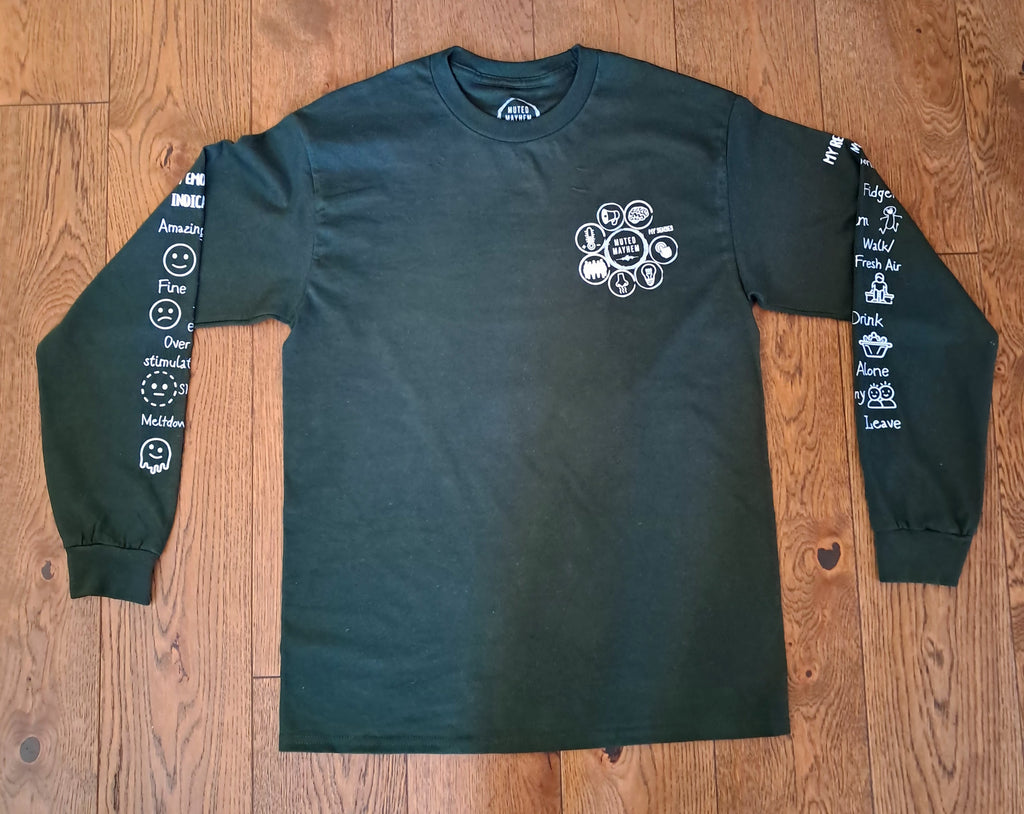 A dark forest green long-sleeve t-shirt with white graphic prints, laid flat on a wooden floor. The shirt features a circular graphic of military-style insignia on the left chest and white text and symbols printed down both sleeves.