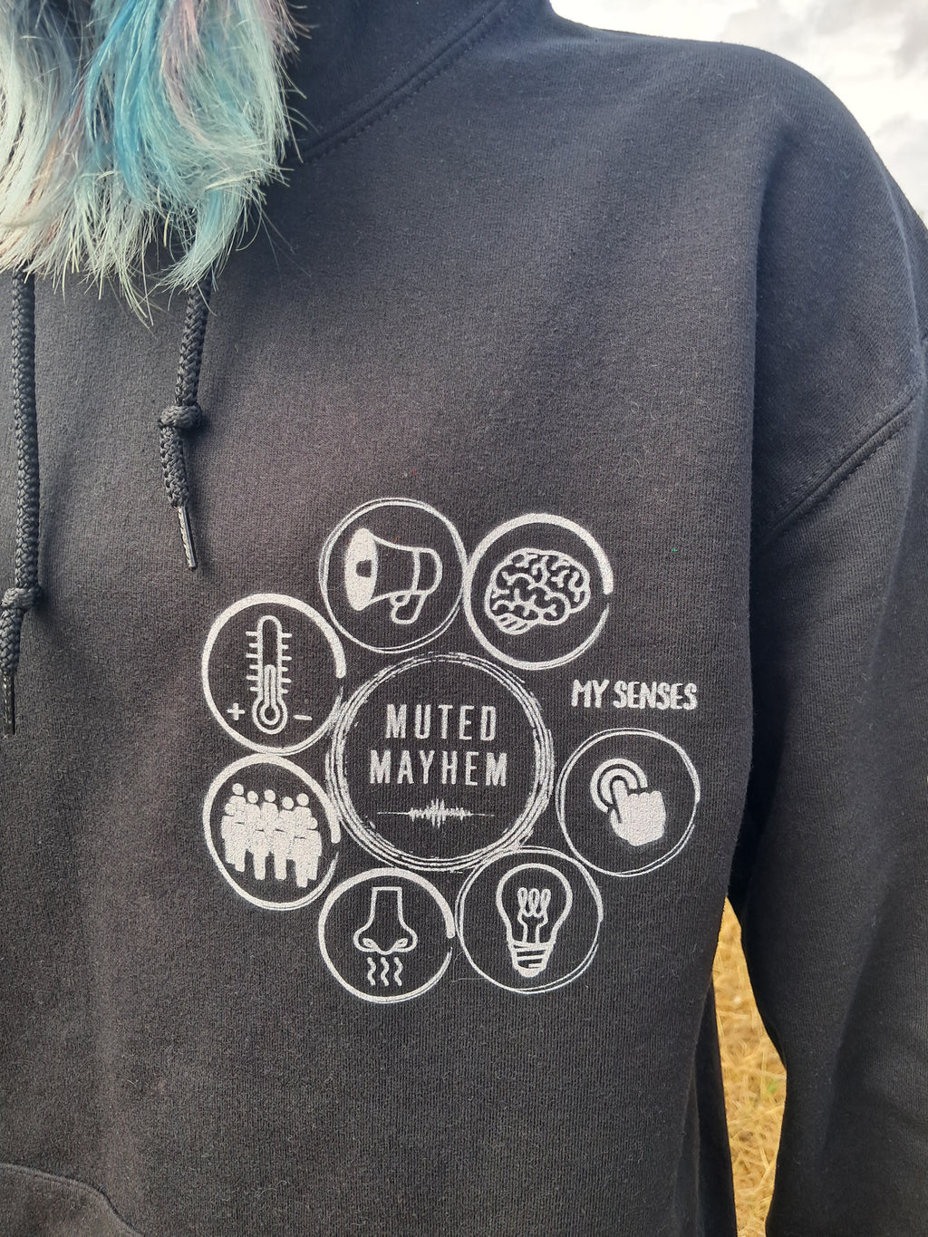 A close-up of a person wearing a dark charcoal gray hoodie with a white graphic print on the chest. The graphic features the text 'MUTED MAYHEM' in the center surrounded by eight circular icons representing senses (temperature, sound, brain, hand, crowd, nose, light bulb, and a megaphone).