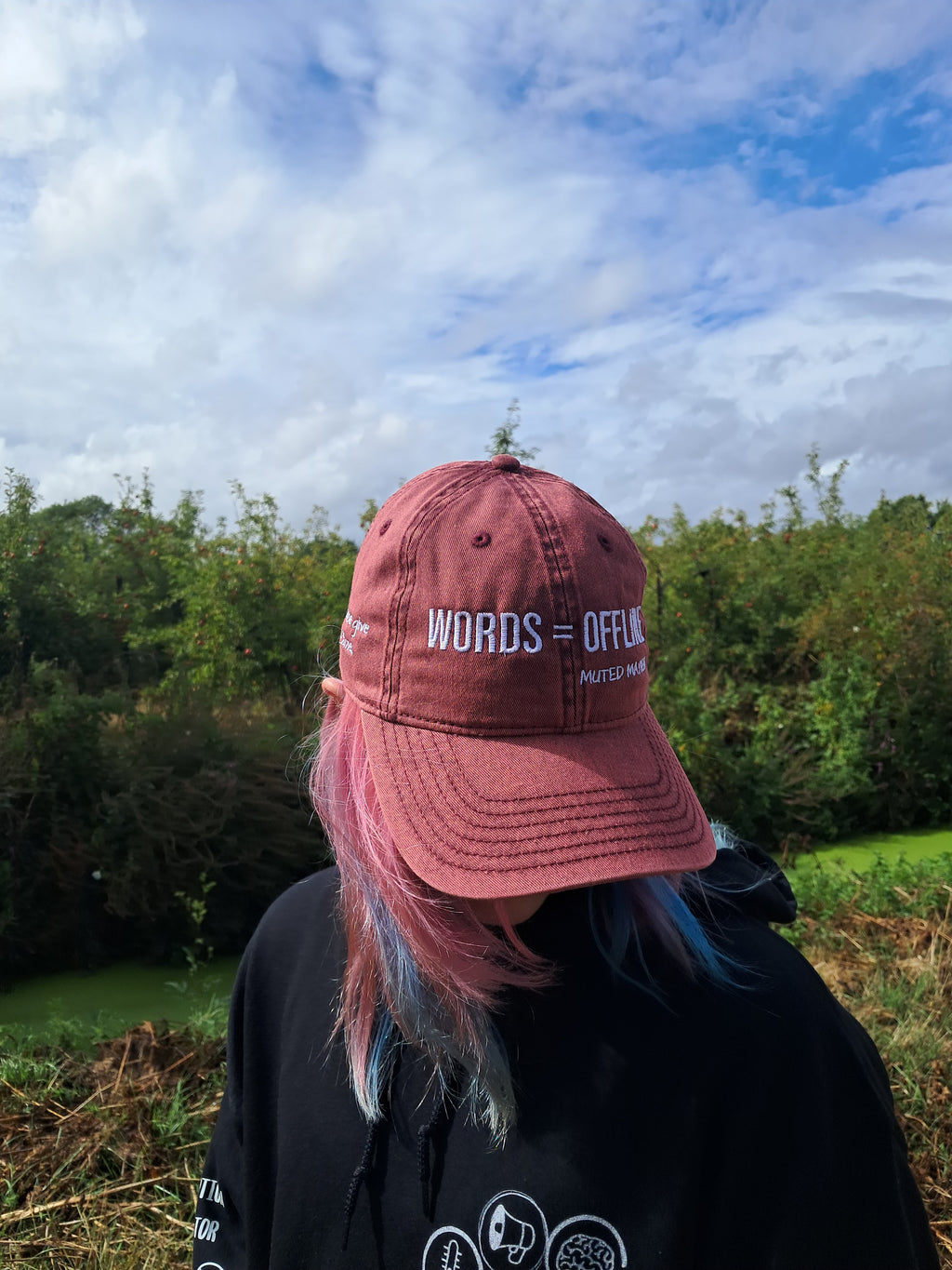 A person outdoors wearing a dusty rose baseball cap with 'WORDS = OFFENSE' printed in white on the front, paired with a black hoodie. The background features green foliage and a blue sky with white clouds.