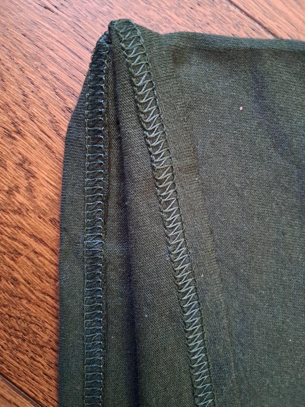Close-up of a dark olive green garment, showing a close-up of a zigzag stitched seam on a wooden surface.