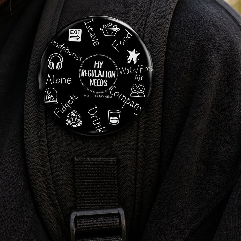 A round black pin badge with white text and icons, featuring the message 'MY REGULATION NEEDS' and surrounding phrases like 'Leave', 'Food', 'Walk/Fresh Air', 'Company', and 'Drink', worn on a black backpack strap.