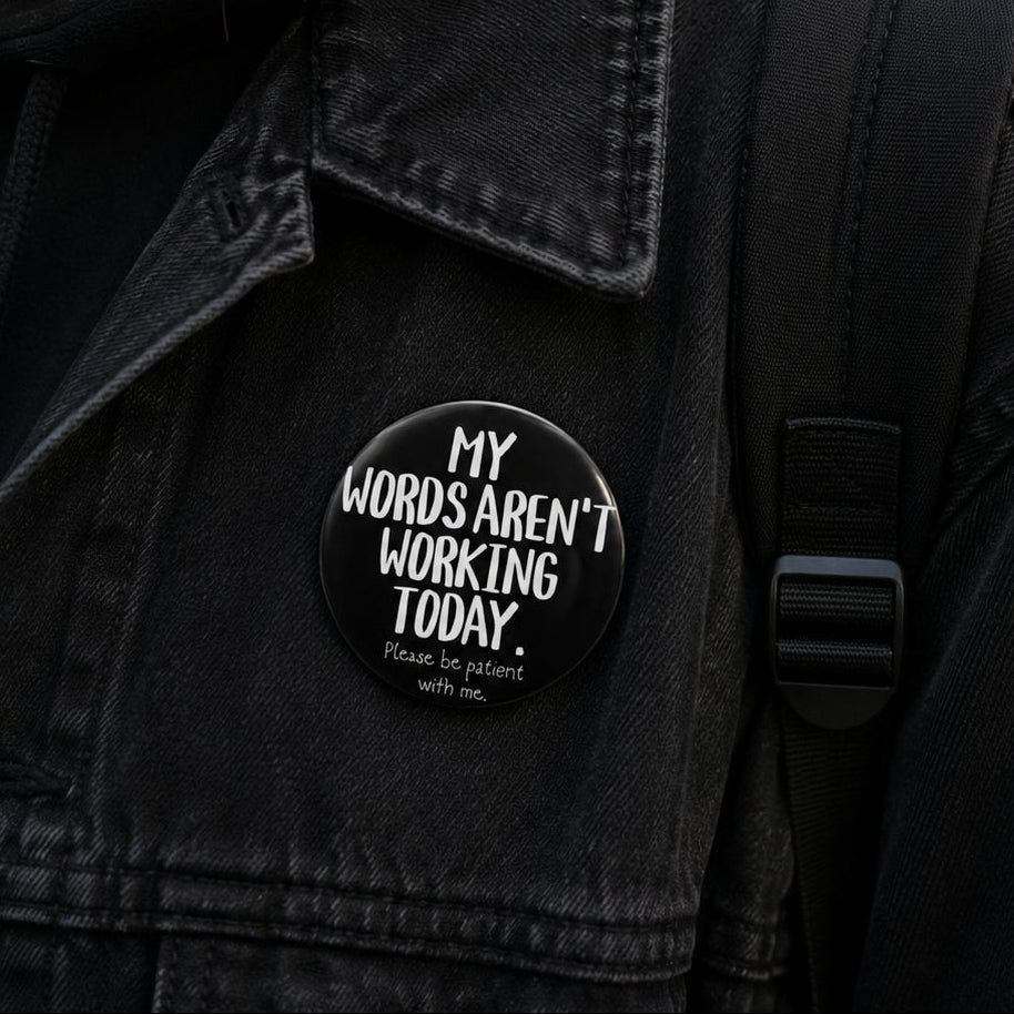 A close-up of a person wearing a black denim jacket and black hoodie, featuring a round black button pin with the text 'MY WORDS AREN'T WORKING TODAY. Please be patient with me.