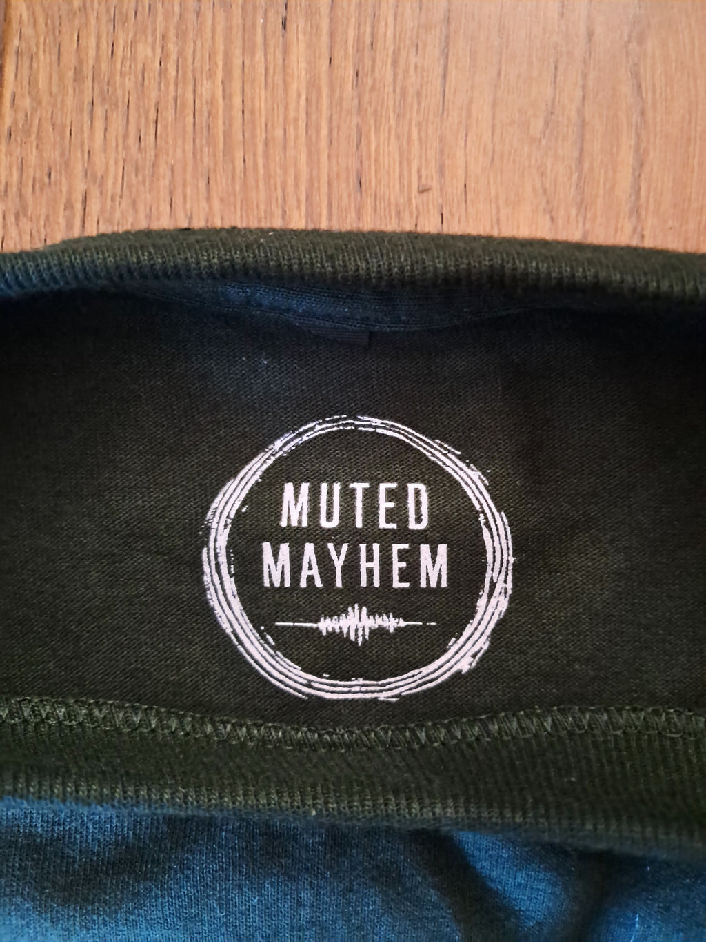 Close-up of a dark olive green t-shirt collar with a white circular 'Muted Mayhem' logo printed inside the neck, laid flat on a wooden surface.