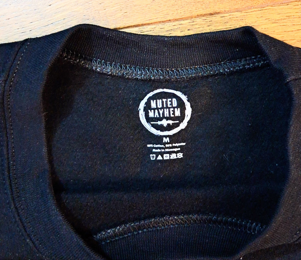 Close-up of the inside collar of a black crewneck t-shirt on a light wood surface, showing the white printed 'Muted Mayhem' brand logo.