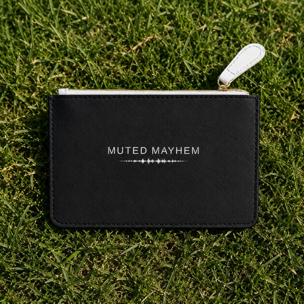 A black textured zip pouch with a white leather pull tab and white printed text reading 'MUTED MAYHEM' lies on green grass.