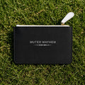 A black textured zip pouch with a white leather pull tab and white printed text reading 'MUTED MAYHEM' lies on green grass.