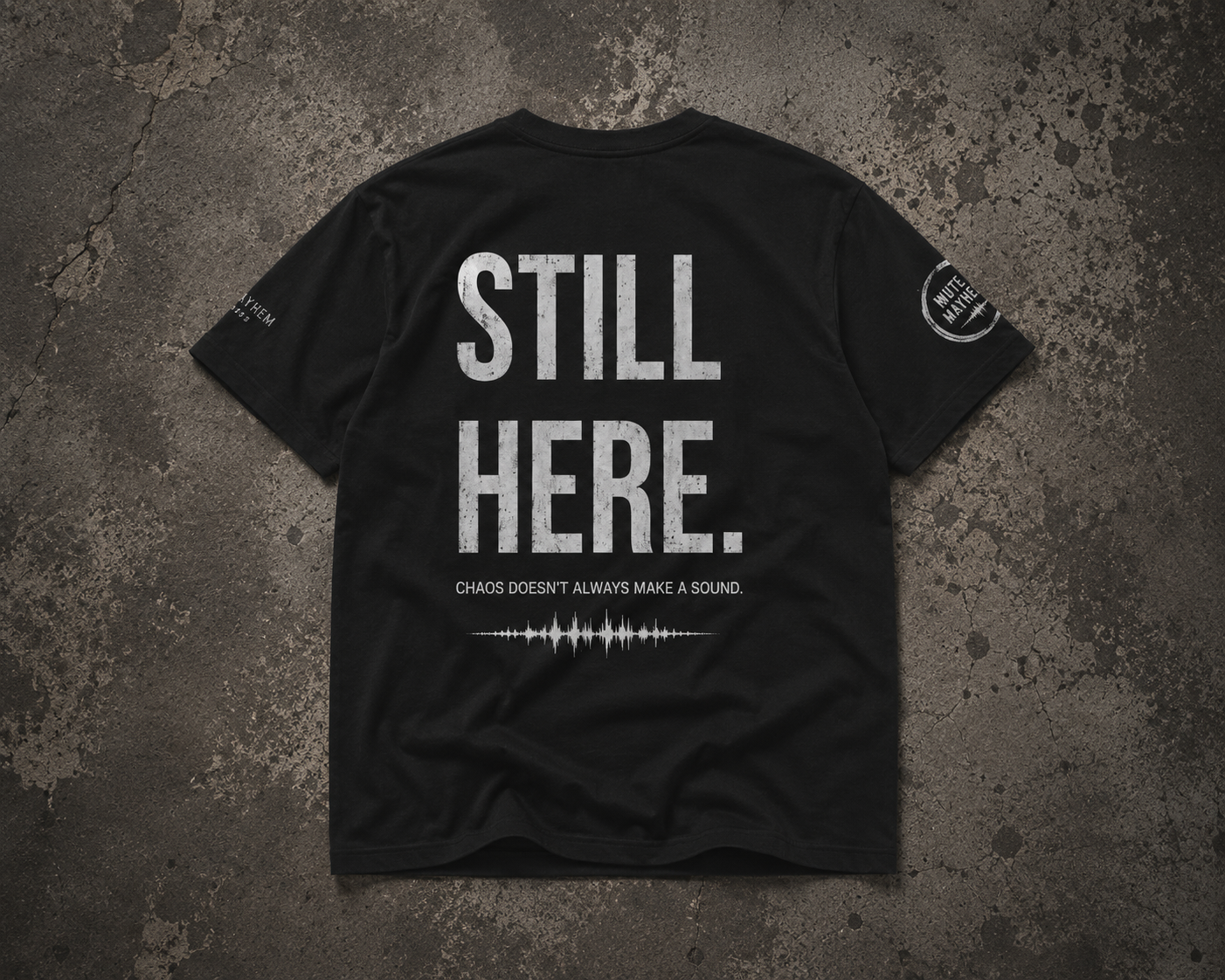 A black short-sleeve t-shirt shown from the back, laid flat on a textured dark gray concrete-style surface. The shirt features large distressed white text reading 'STILL HERE.' promoting mental health and neurodivergence awareness by muted mayhem.