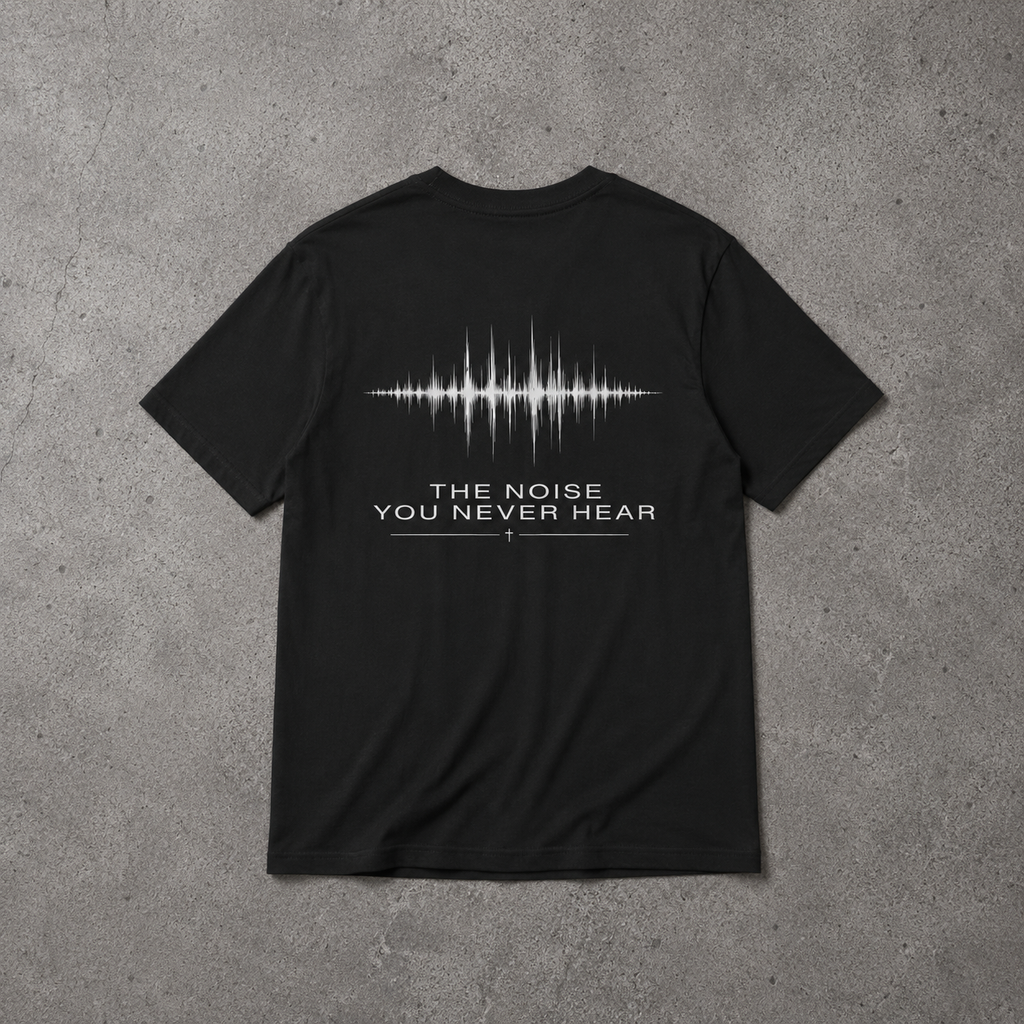 A black short-sleeve t-shirt laid flat on a textured gray concrete background, showing the back side with a white soundwave graphic and the text 'THE NOISE YOU NEVER HEAR' promoting mental health and neurodivergence awareness by muted mayhem.