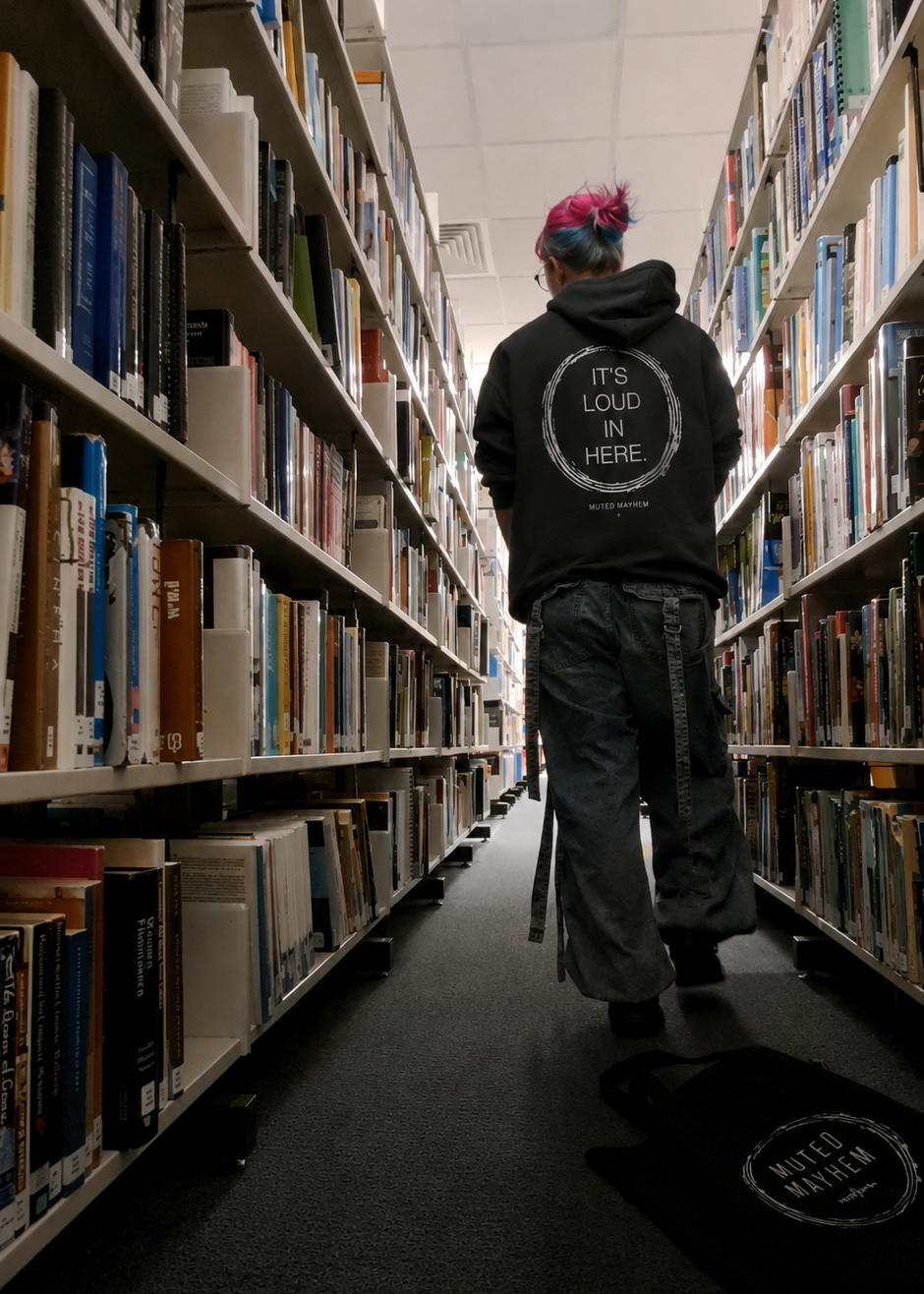 A person viewed from behind walking down a narrow aisle lined with bookshelves, wearing a black hoodie with a circular white graphic that reads 'THE LOUD IN HERE' and dark pants. The setting is a library or bookstore with rows of books on shelves promoting mental health and neurodivergence awareness by muted mayhem